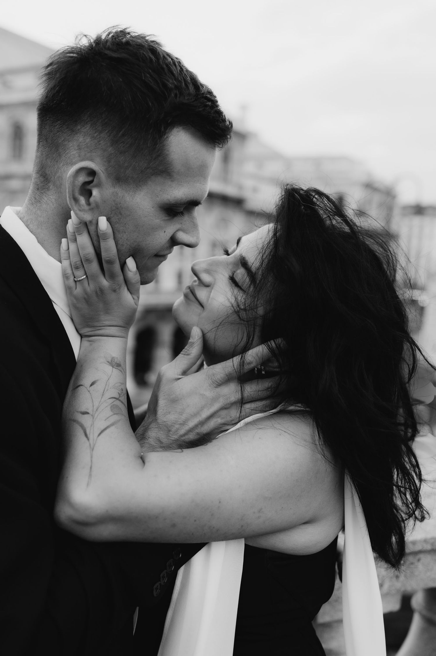 Black and white photo of a romantic couple almost kissing, woman with dark hair holding mans face tenderly.