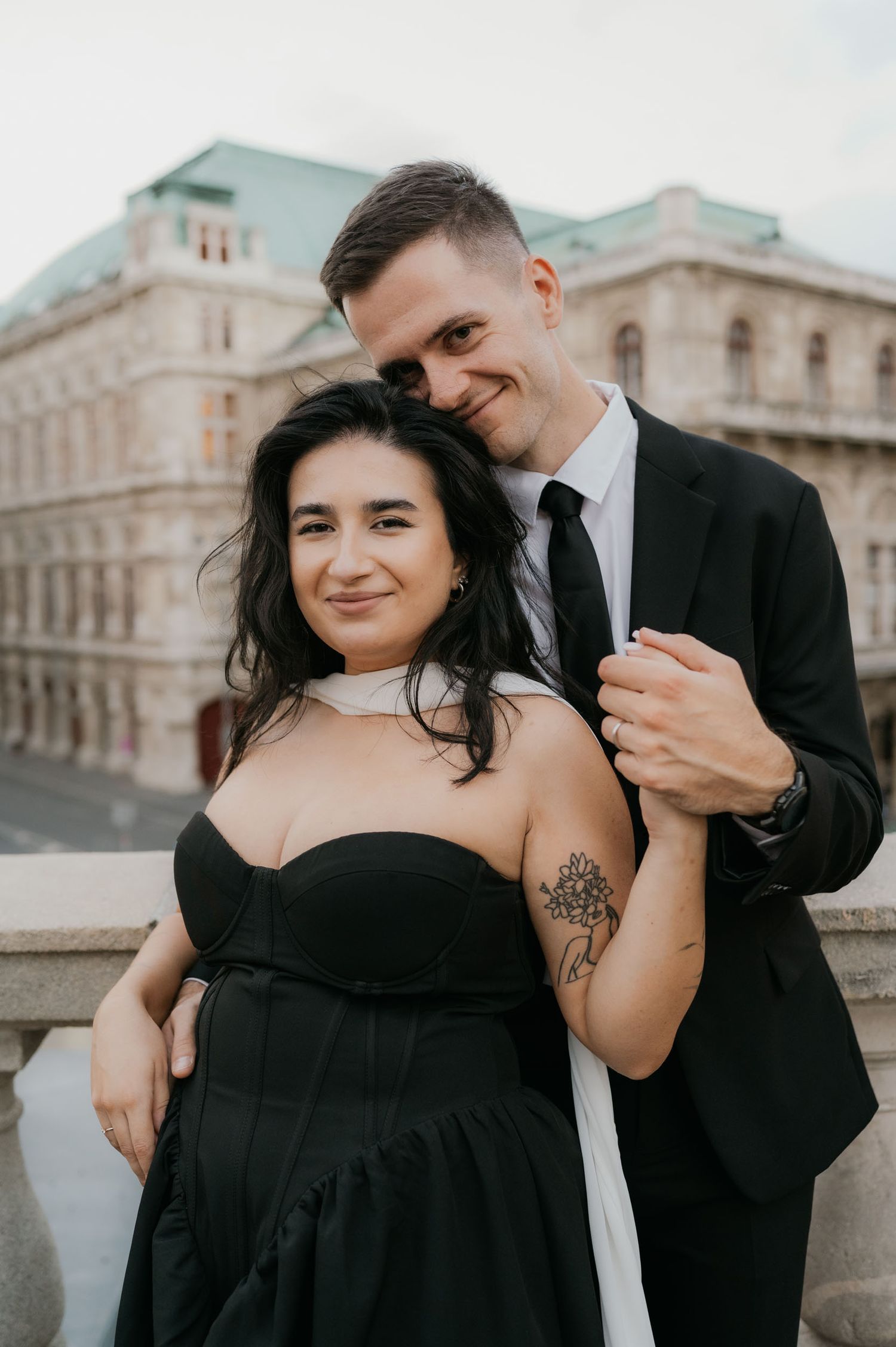 Couple in formal attire embracing outdoors, woman in black strapless dress with floral tattoo, man in black suit.