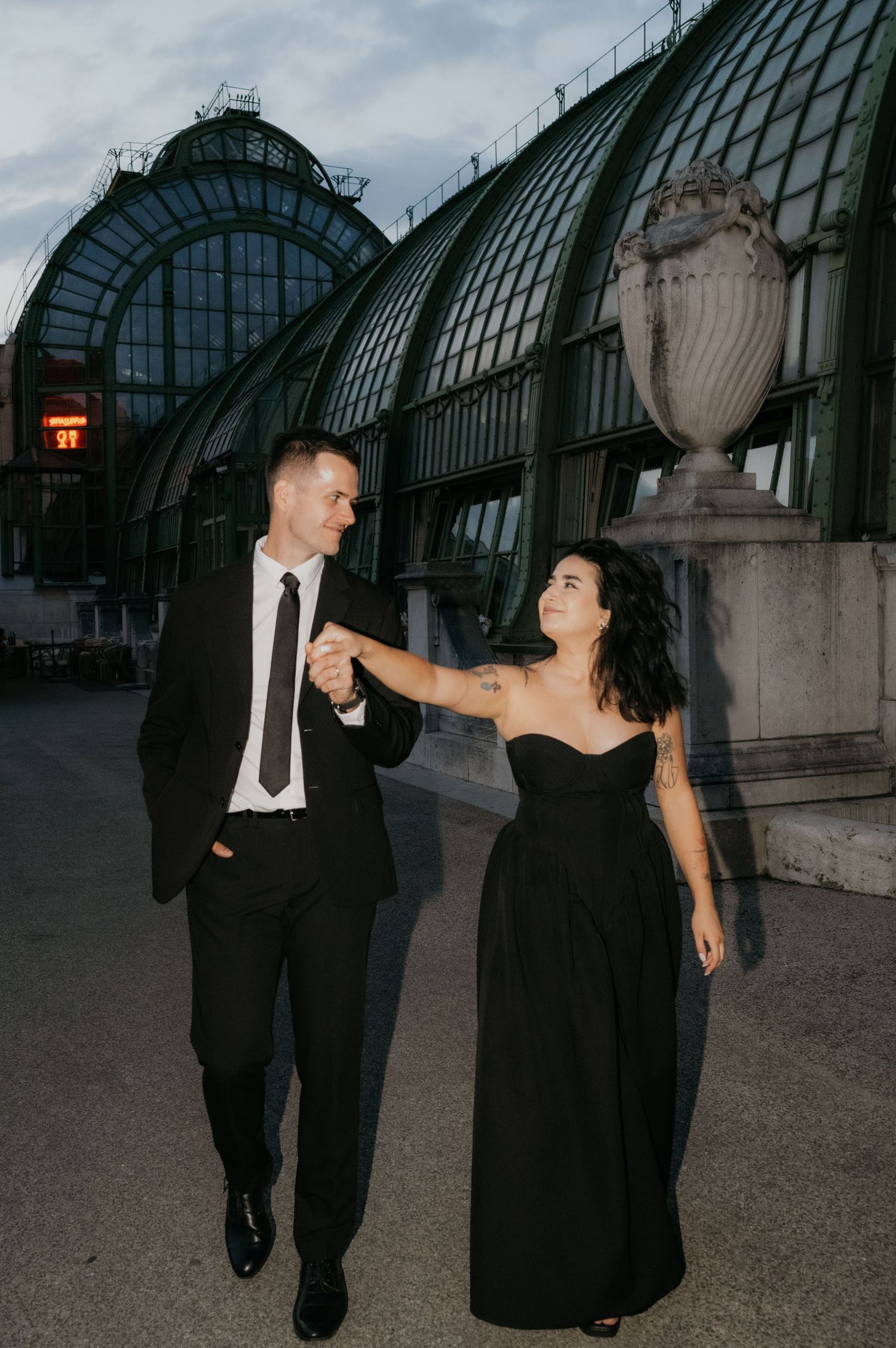 Elegant couple in matching black outfits holding hands outside a grand glass greenhouse at dusk.