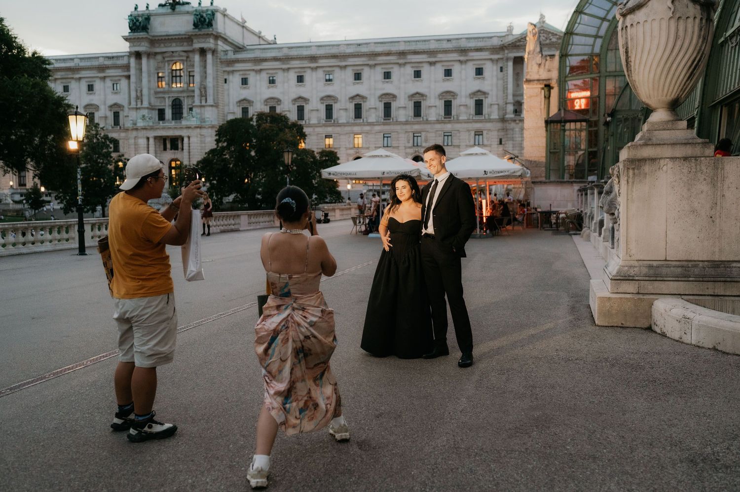 Couple in formal black attire poses for photos in front of a grand European palace at dusk.