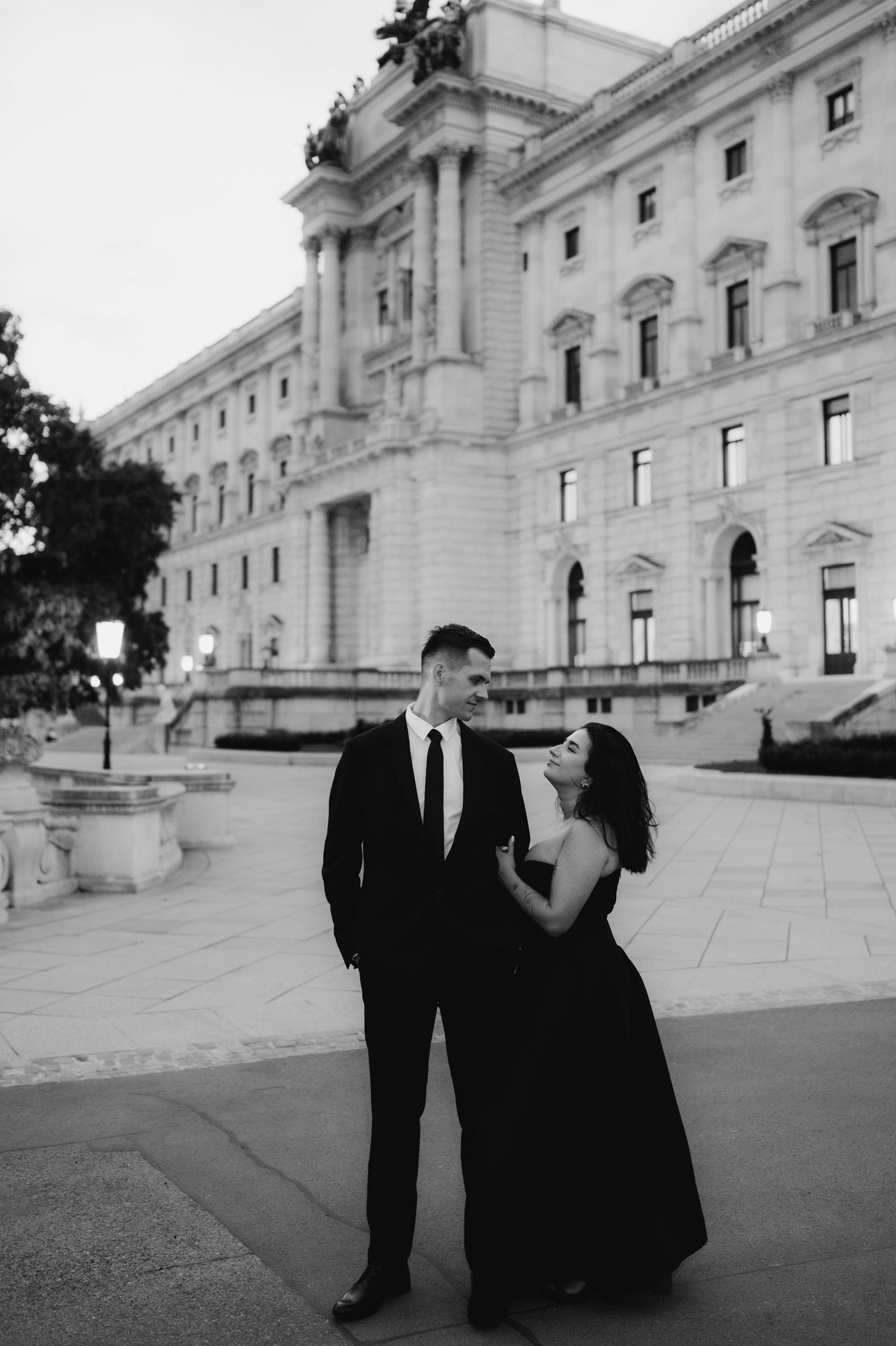 Elegant couple in black formal attire posing in front of a grand neoclassical building in a black and white photo.
