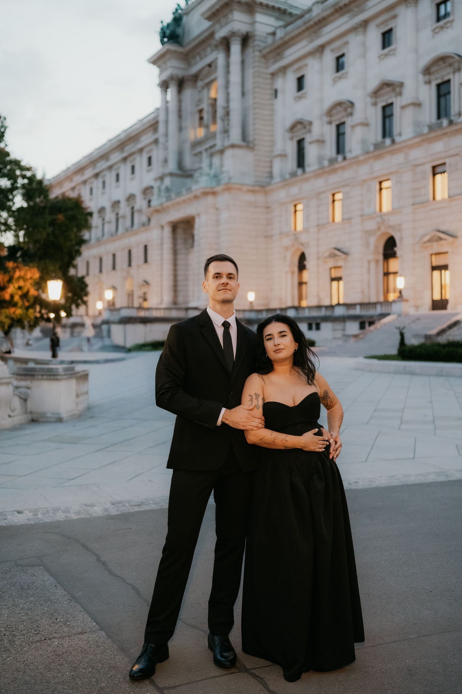 Elegant couple in matching black formal attire posing in front of a grand neoclassical building at dusk.