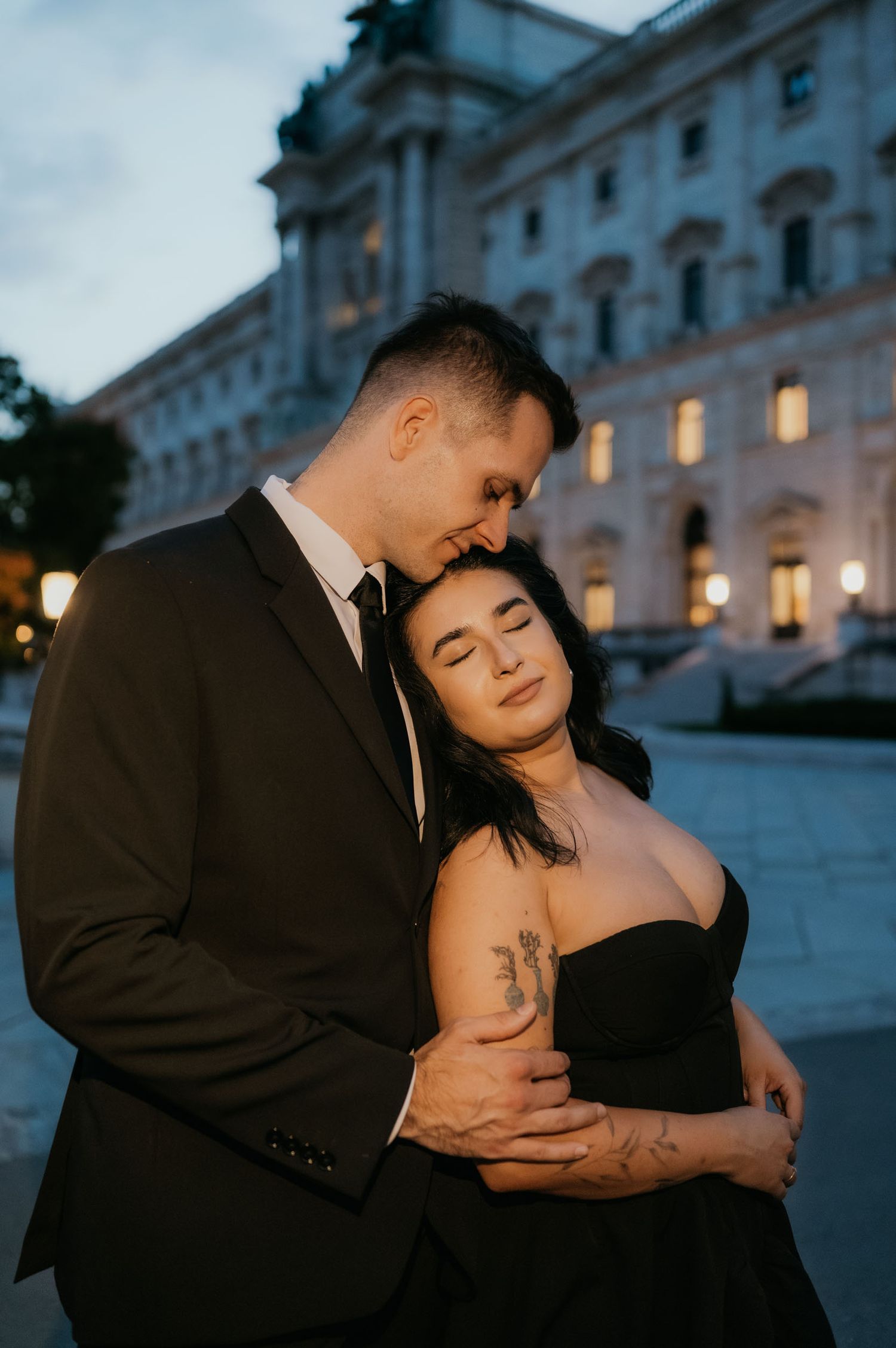 Couple in black formal wear embrace at dusk outside a grand building, woman with tattoos leans back into mans arms.