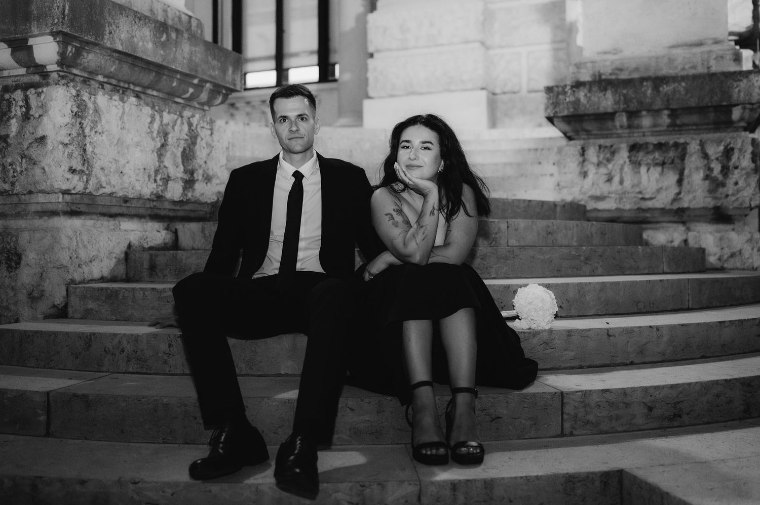 Black and white photo of an elegant couple sitting on stone steps, man in suit and woman in black dress.