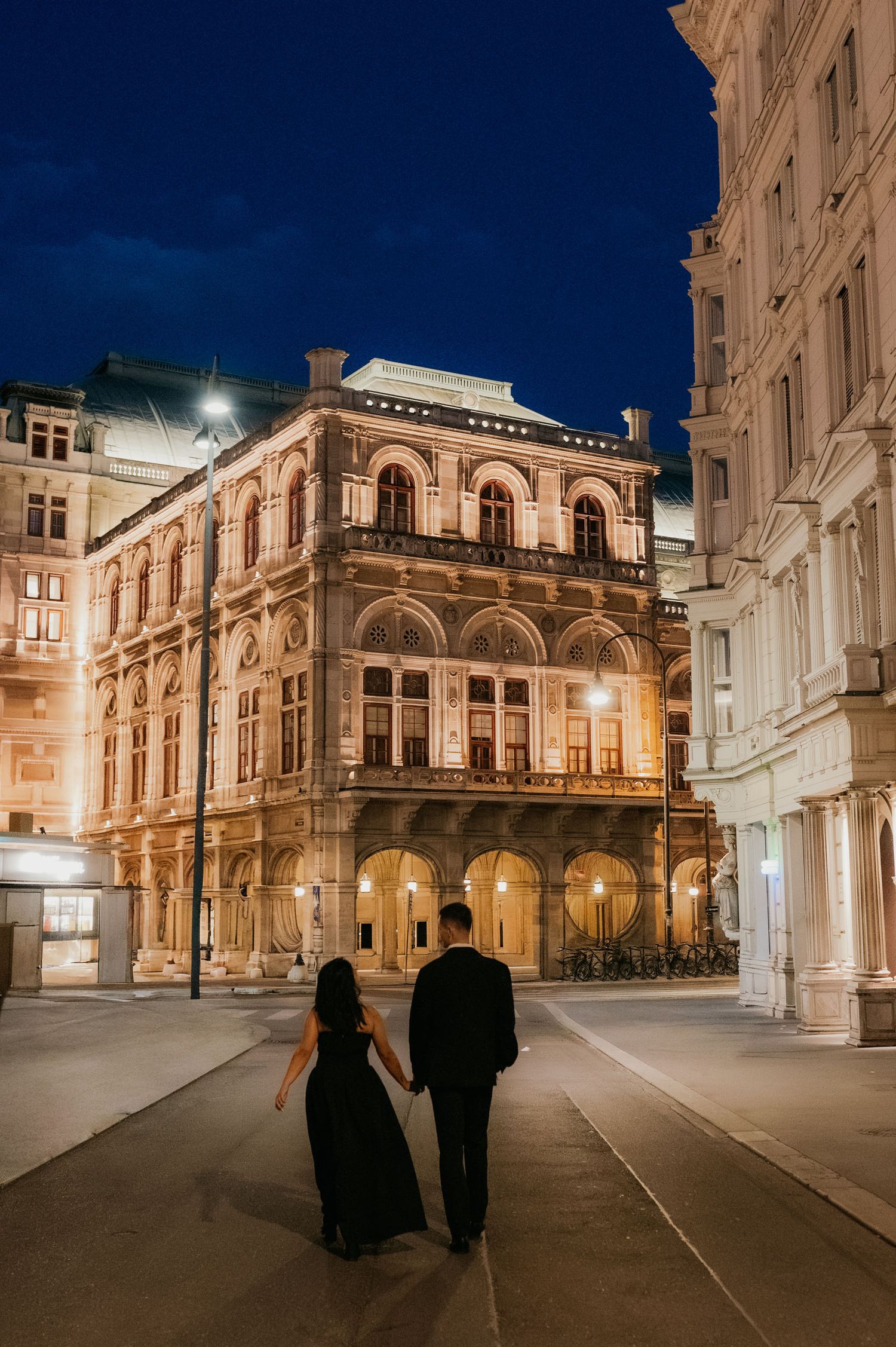 Couple holding hands walking toward illuminated historic European building at night in elegant formal attire.