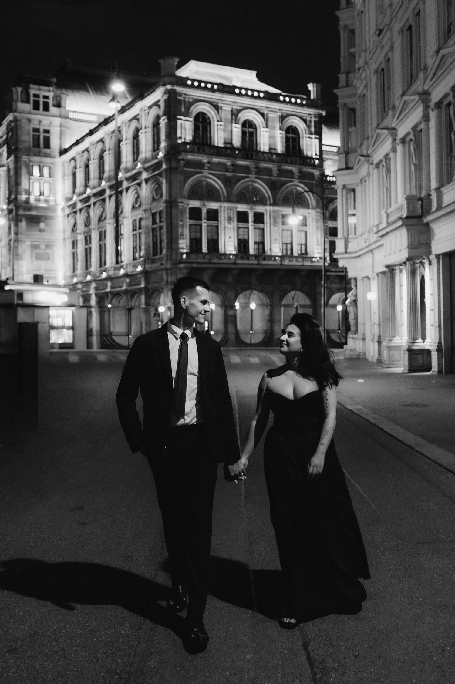Black and white photo of an elegant couple holding hands walking at night in front of a grand historic building.