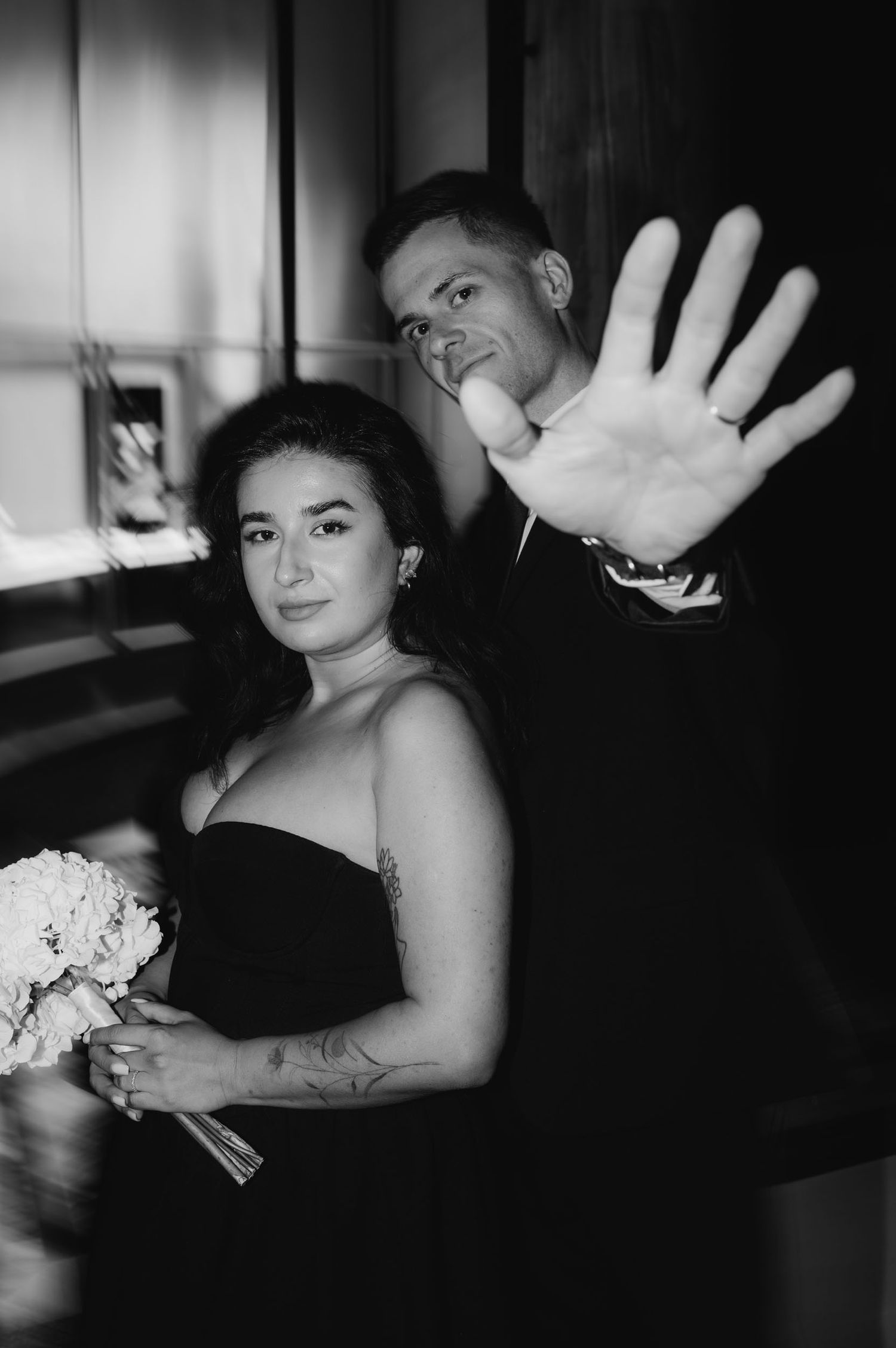 Black and white photo of a couple, woman in strapless dress holding bouquet, man raising hand to camera.