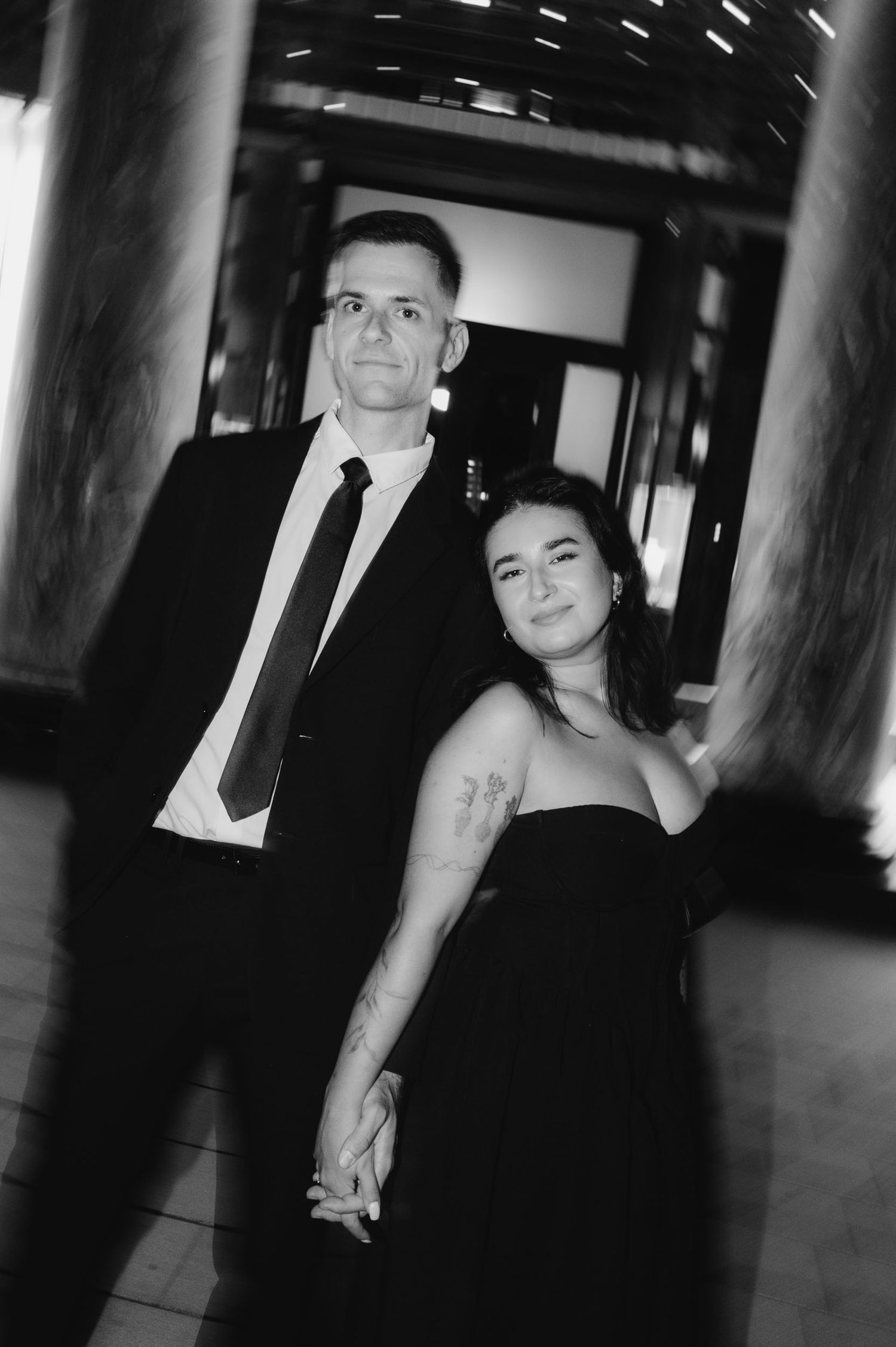 Black and white photo of a couple holding hands, man in suit and tie, woman in strapless dress with tattoo, elegant venue.
