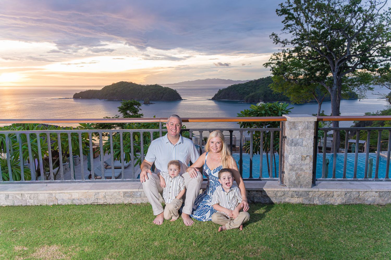 Happy family of three posing on a beach at sunset, with parents smiling and toddler pointing.