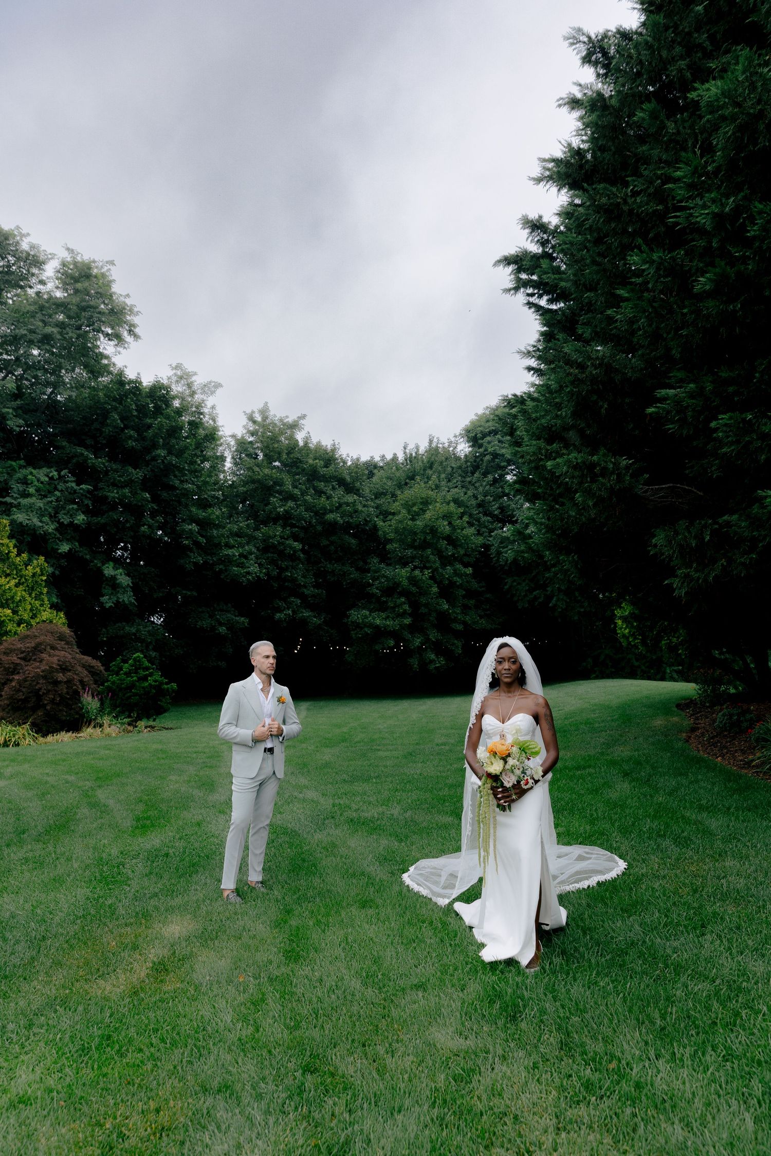 A wedding couple poses on a lush green lawn surrounded by tall trees on an overcast day.