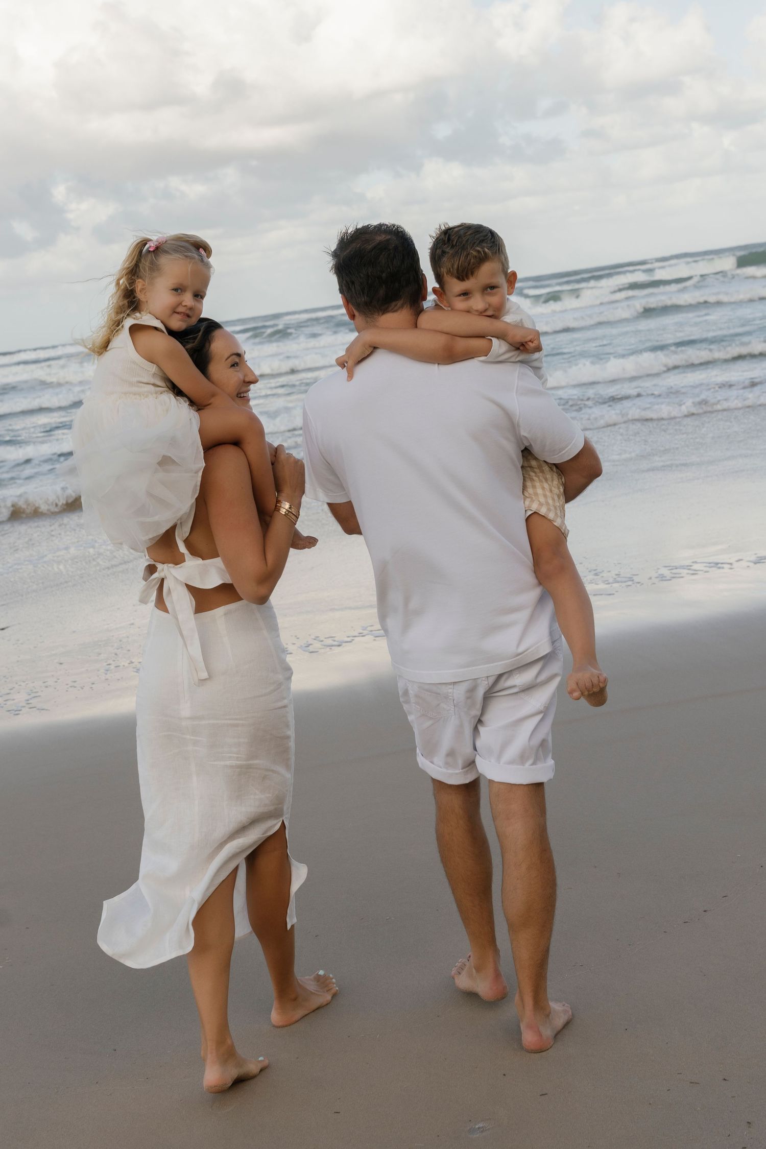 Family in white outfits enjoying a beach walk, parents carrying children on their backs near ocean waves.
