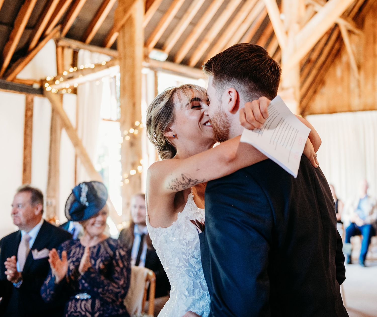 Bride and groom share first dance embrace in rustic barn venue with fairy lights and guests applauding in background.