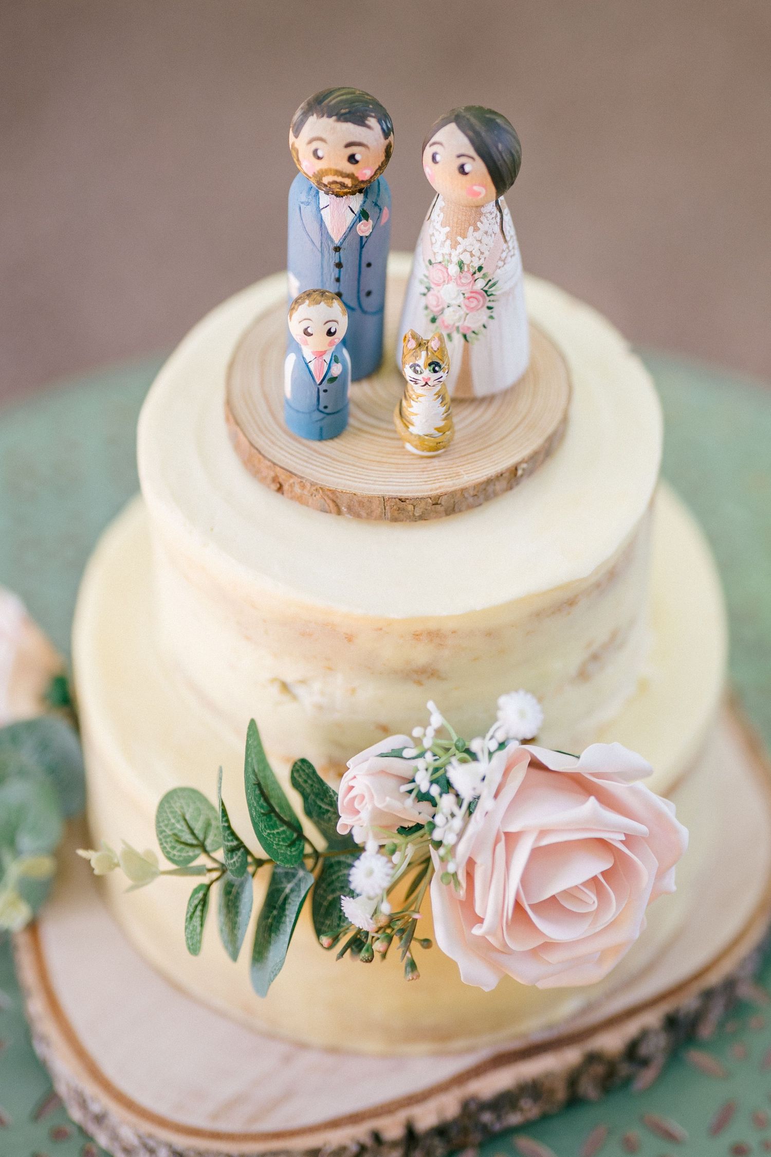 Two-tier semi-naked wedding cake with wooden slice base, pink roses, and custom family cake toppers.