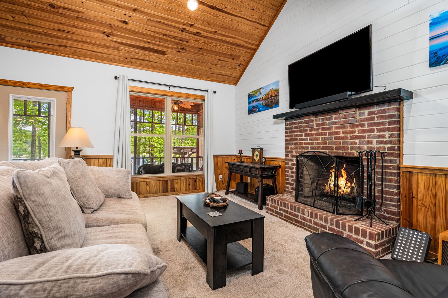 Cozy cabin living room with brick fireplace, wood-paneled walls, vaulted ceiling, mounted TV, and plush seating on carpet flooring.