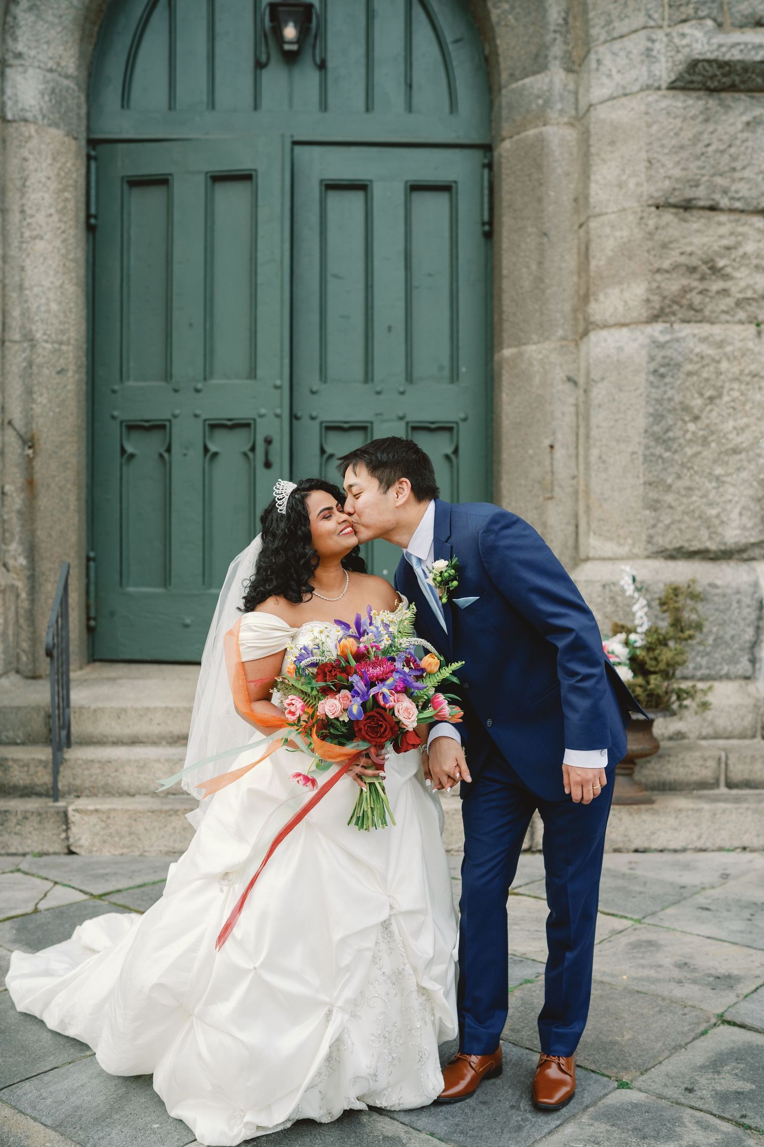 Bride and groom share a kiss outside a green wooden church door, bride holding a vibrant colorful bouquet.