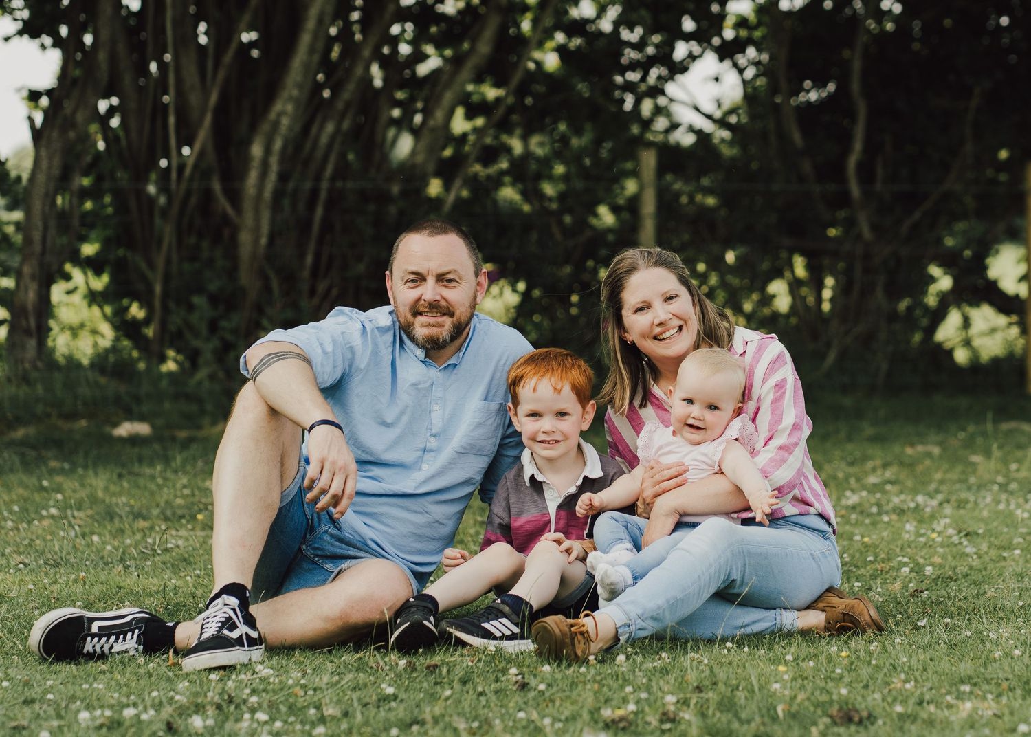 Happy family of four sitting on green grass outdoors, with two young children, surrounded by lush trees on a sunny day.