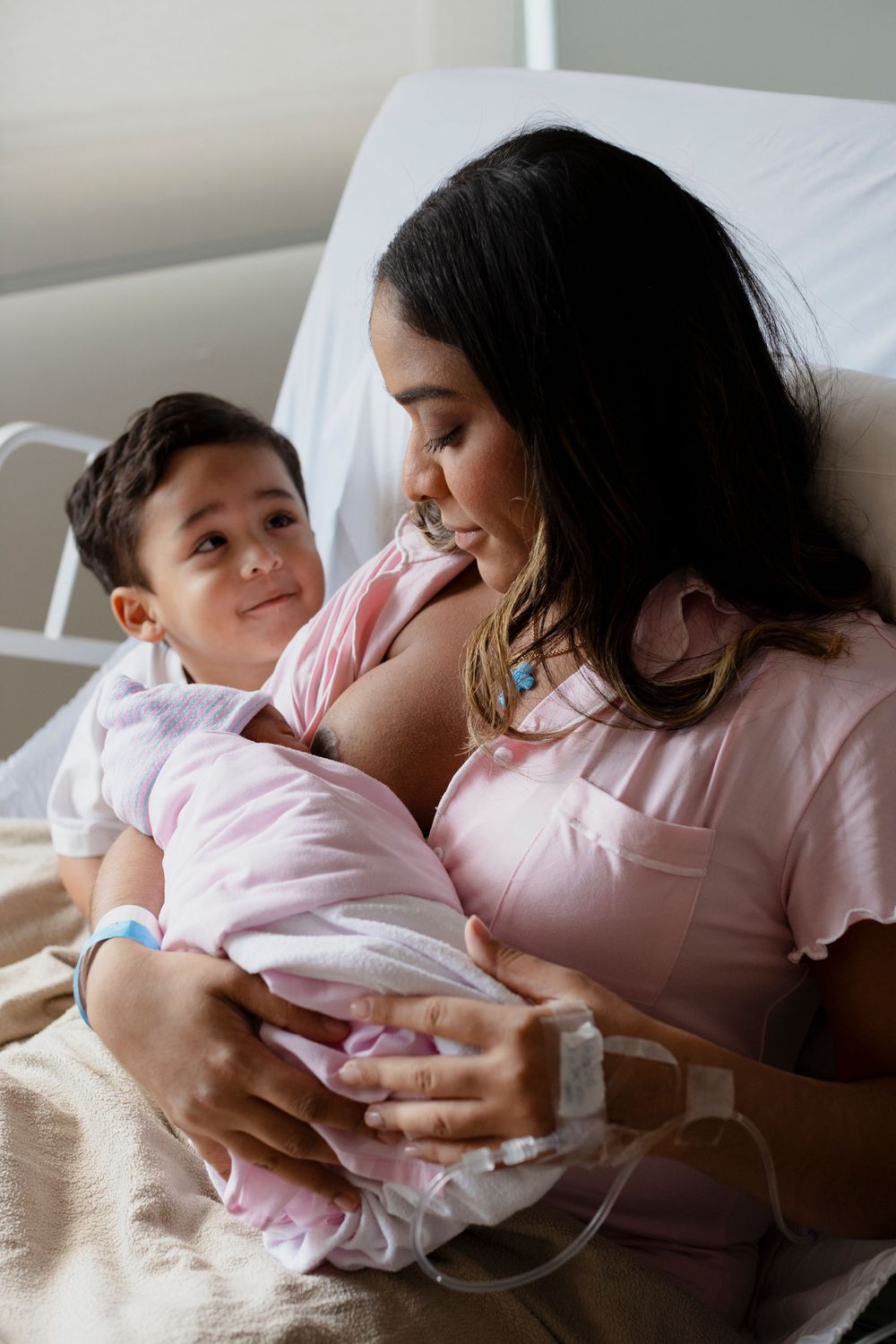 A mother in a hospital bed tenderly holds her newborn baby while an older child sits beside her, all dressed in pink.