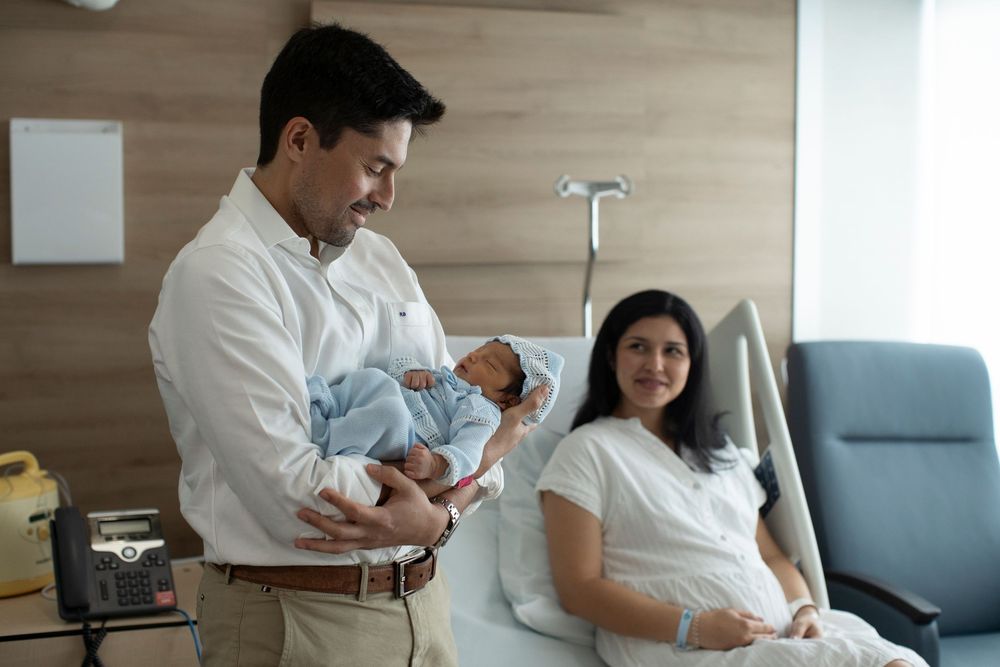 New father holds newborn baby in hospital room while mother rests in bed after childbirth.
