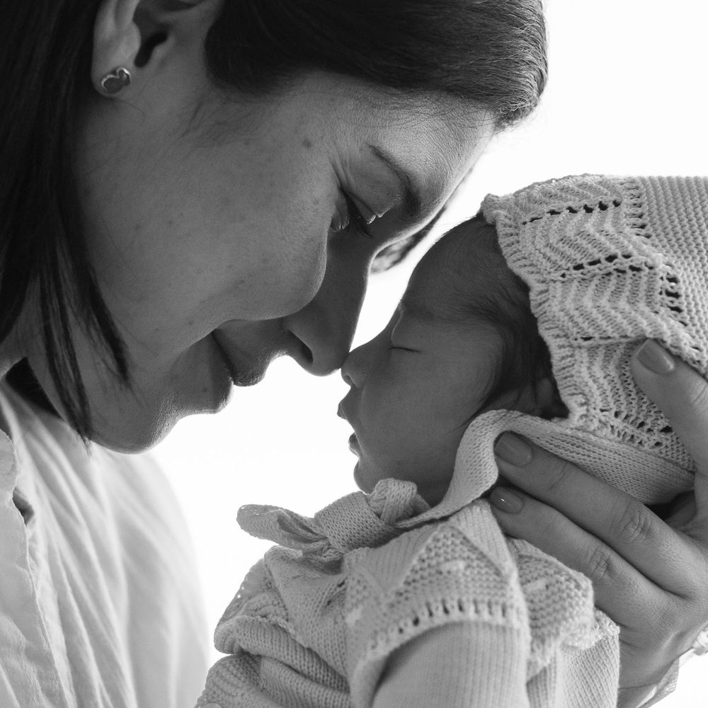 Black and white photo of a mother tenderly touching foreheads with her newborn baby wrapped in a knit blanket.