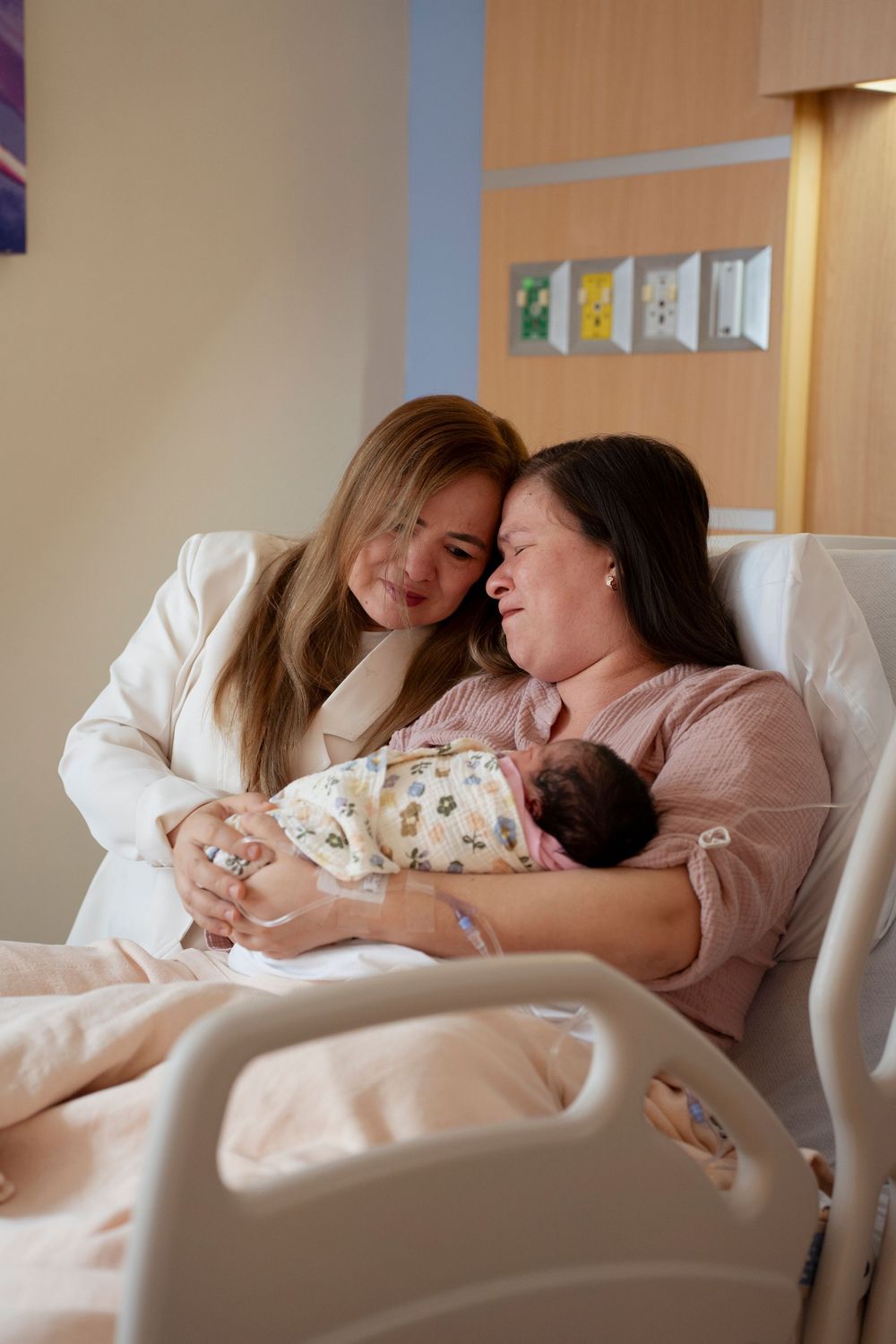 A new mother holds her newborn in a hospital bed while a woman in white leans in to share a tender moment.