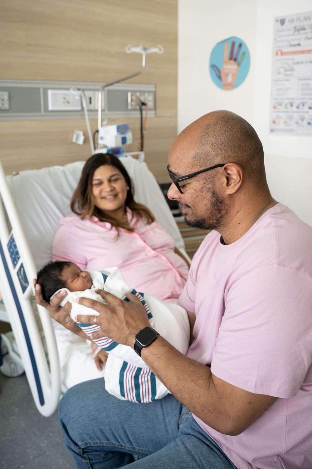 Father holds newborn baby in hospital room while mother rests in bed, smiling after childbirth.