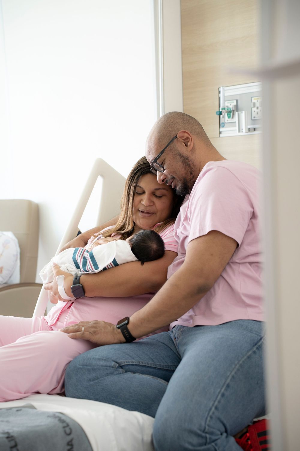New parents sitting on hospital bed lovingly cradling their newborn baby, both wearing pink and jeans.