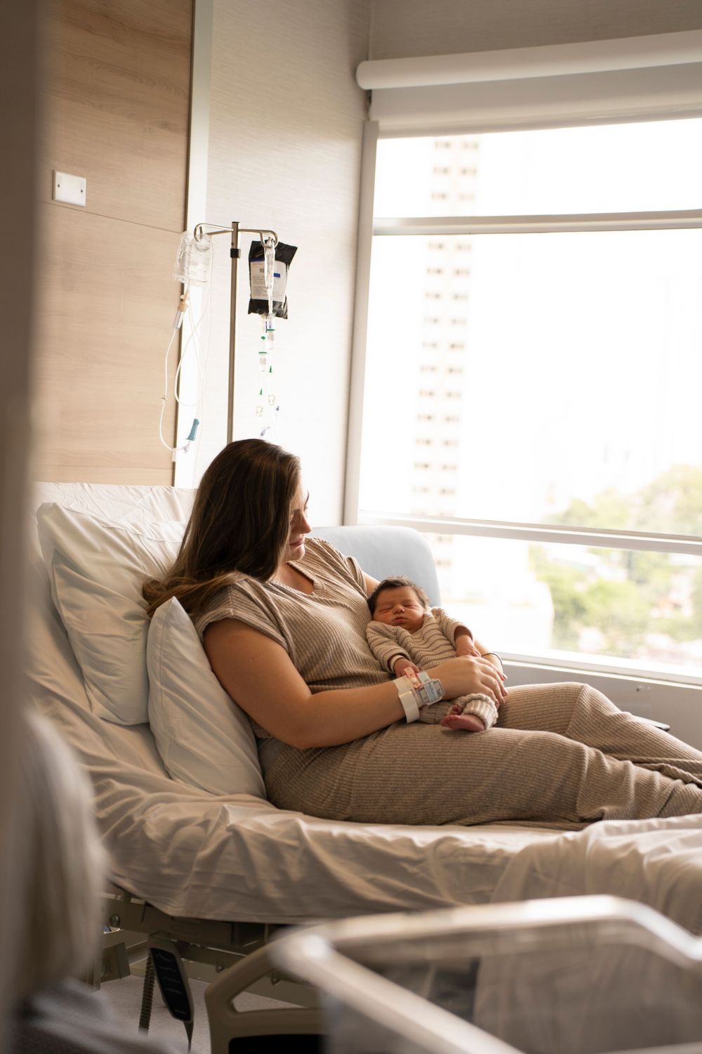 New mother resting in hospital bed holding her newborn baby near a bright window after childbirth.