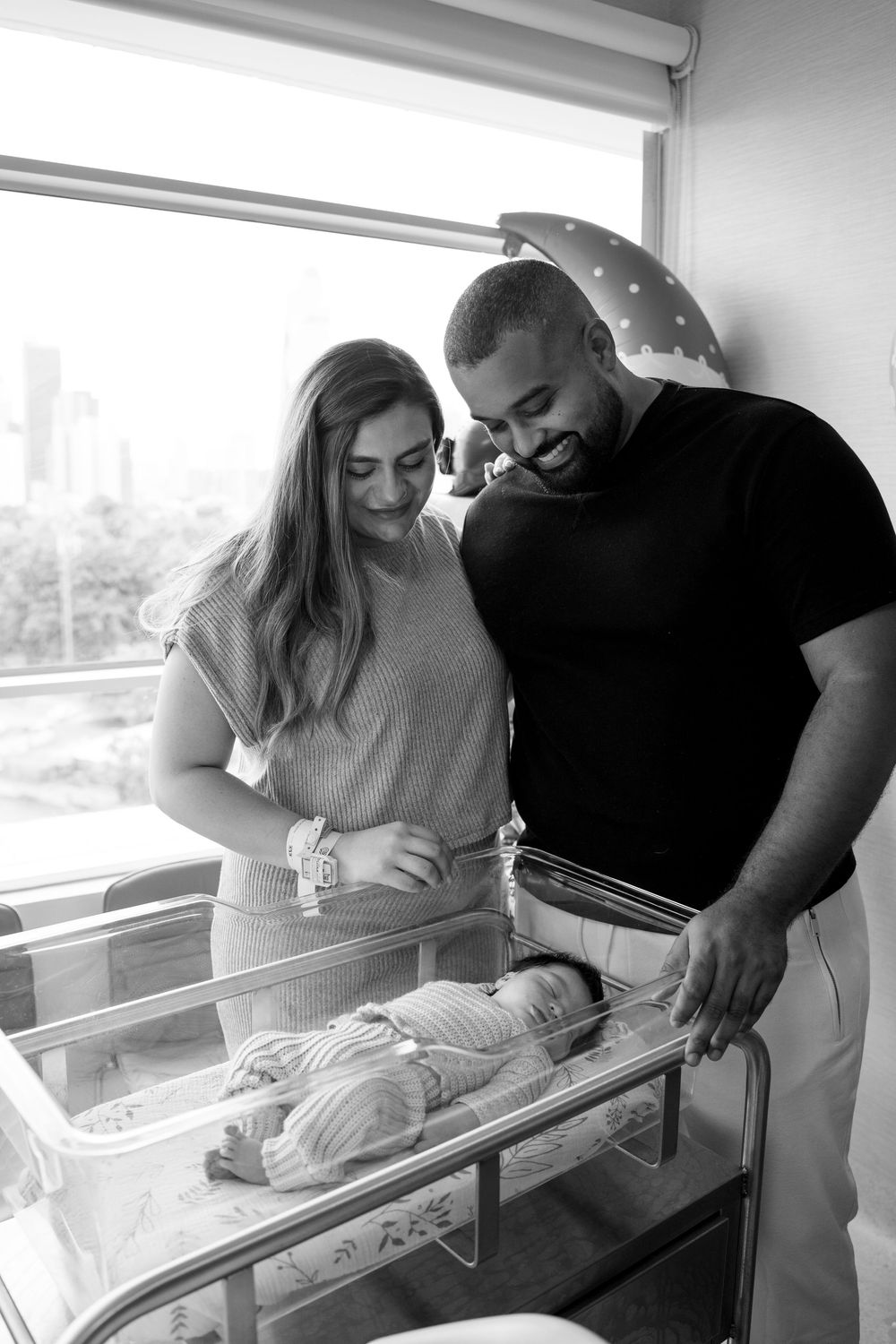 Black and white photo of new parents smiling over their newborn baby in a hospital bassinet.