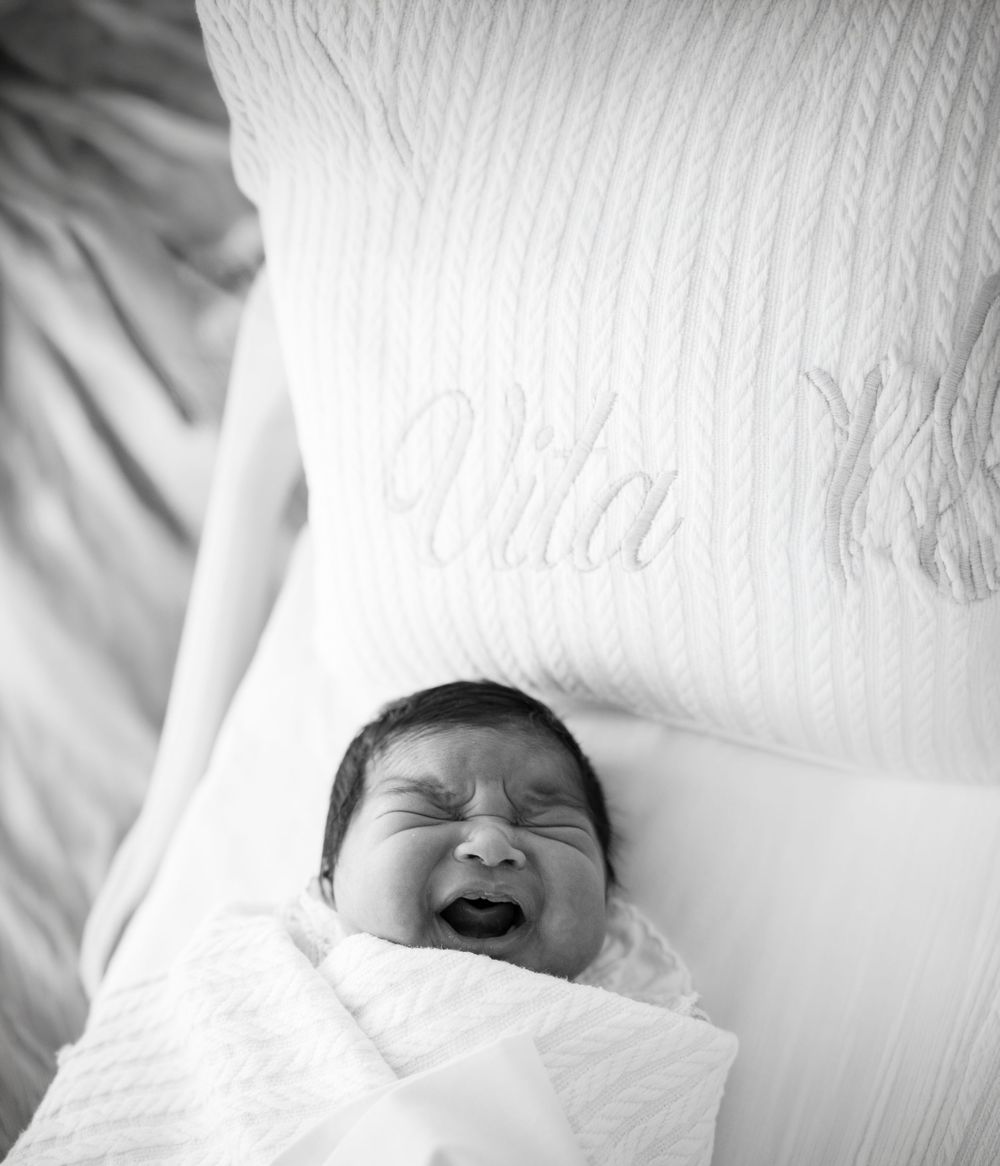 Black and white photo of a yawning newborn baby swaddled in white blanket resting on a pillow embroidered with name Vita.