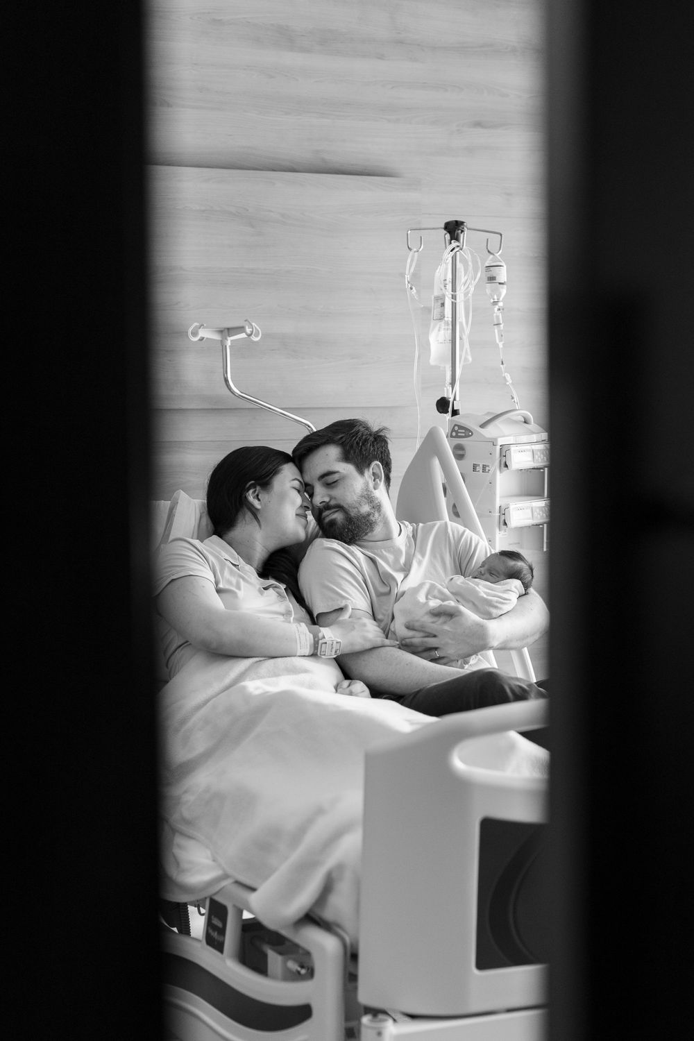 Black and white photo of new parents sharing a tender moment with their newborn baby in a hospital room.