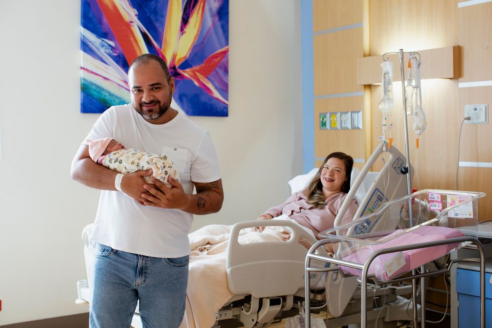 Father holds newborn baby while mother rests in hospital bed after delivery in modern maternity ward.