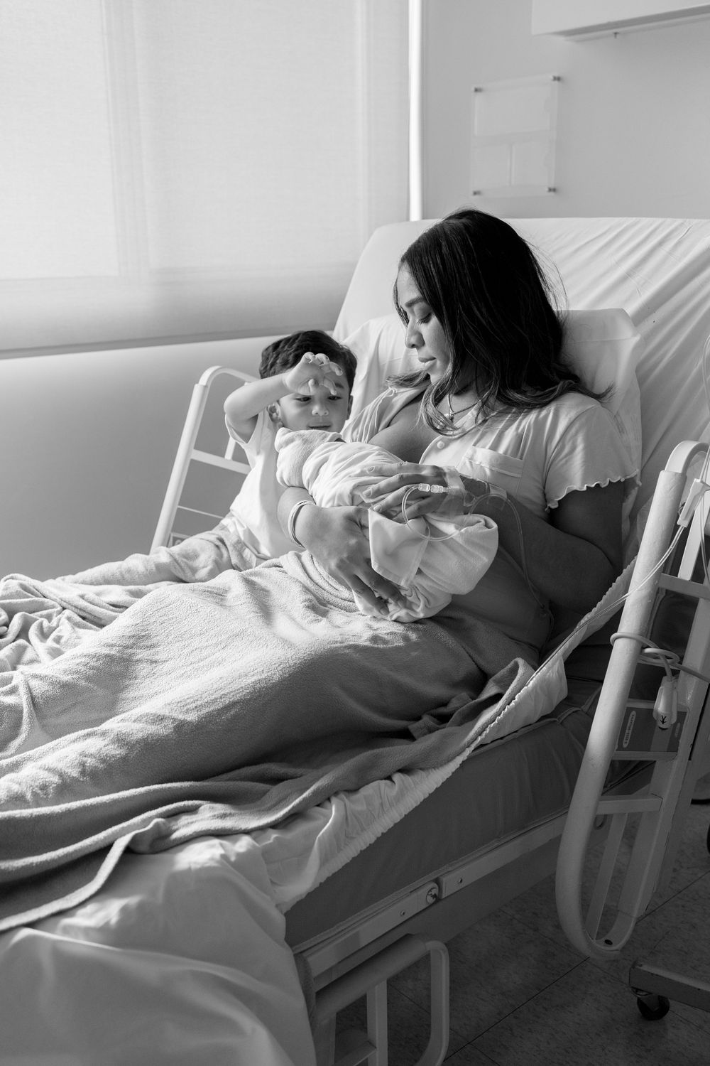 Black and white photo of a mother in hospital bed holding newborn baby with young child beside her.