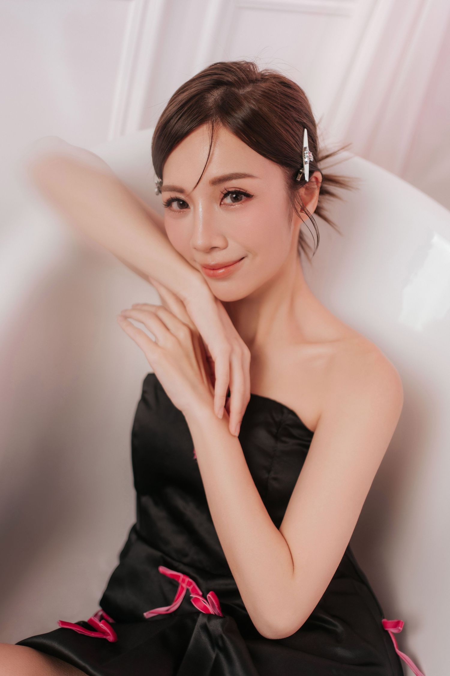 Young woman in black strapless dress with pink accents, hair accessory, posing gracefully on white chair.