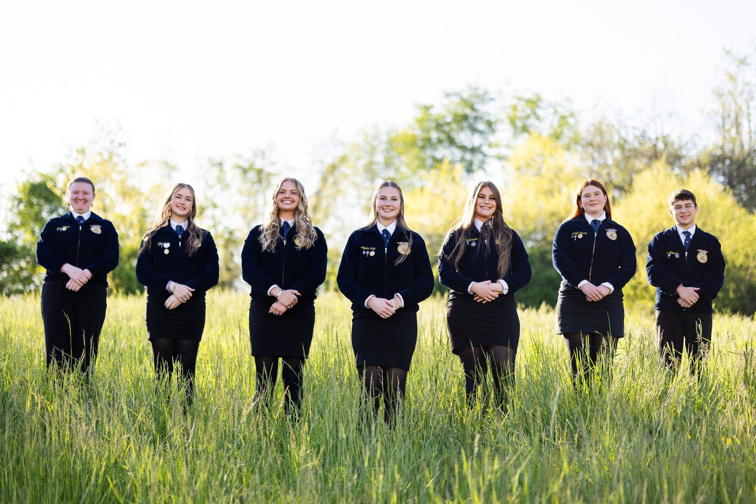 Group of seven students in FFA jackets standing together in a lush green field.