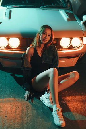 Young Asian woman in denim jacket and black outfit sitting in front of a classic muscle car with glowing headlights at night.