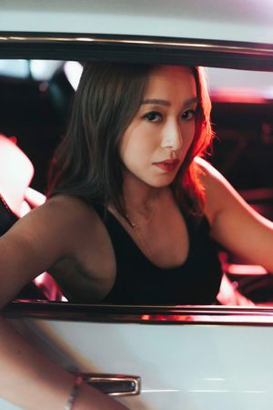 Young woman with long dark hair wearing a black tank top poses inside a car with red ambient lighting.