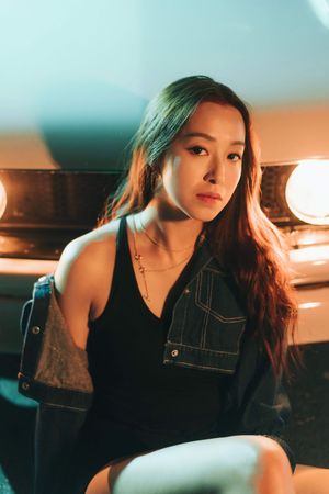 Young Asian woman in denim jacket and black dress posing near vintage car vanity lights in moody teal and orange lighting.
