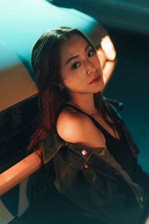 Young Asian woman in denim vest poses under dramatic teal and orange studio lighting.