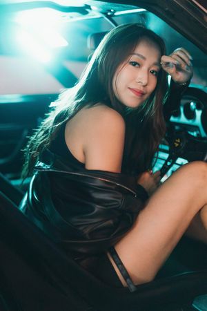 A young Asian woman in a black leather dress poses inside a car with teal lighting.