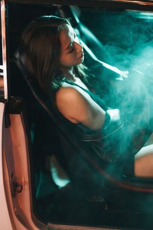 A young woman sits in a car seat surrounded by teal smoke at night, illuminated by dramatic lighting.