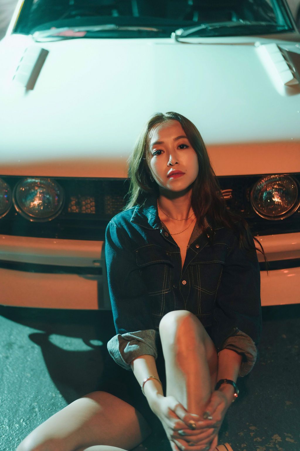 Young Asian woman in denim shirt sitting in front of a classic white muscle car at night with warm lighting.