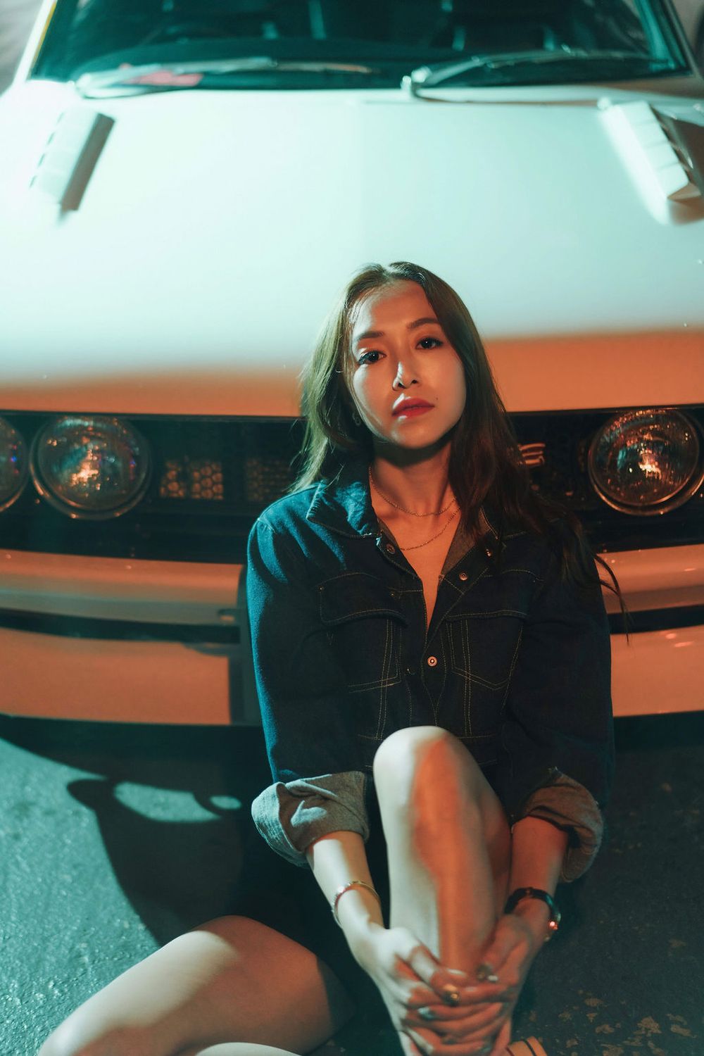 Young Asian woman in denim shirt sitting in front of a classic white muscle car at night with warm lighting.