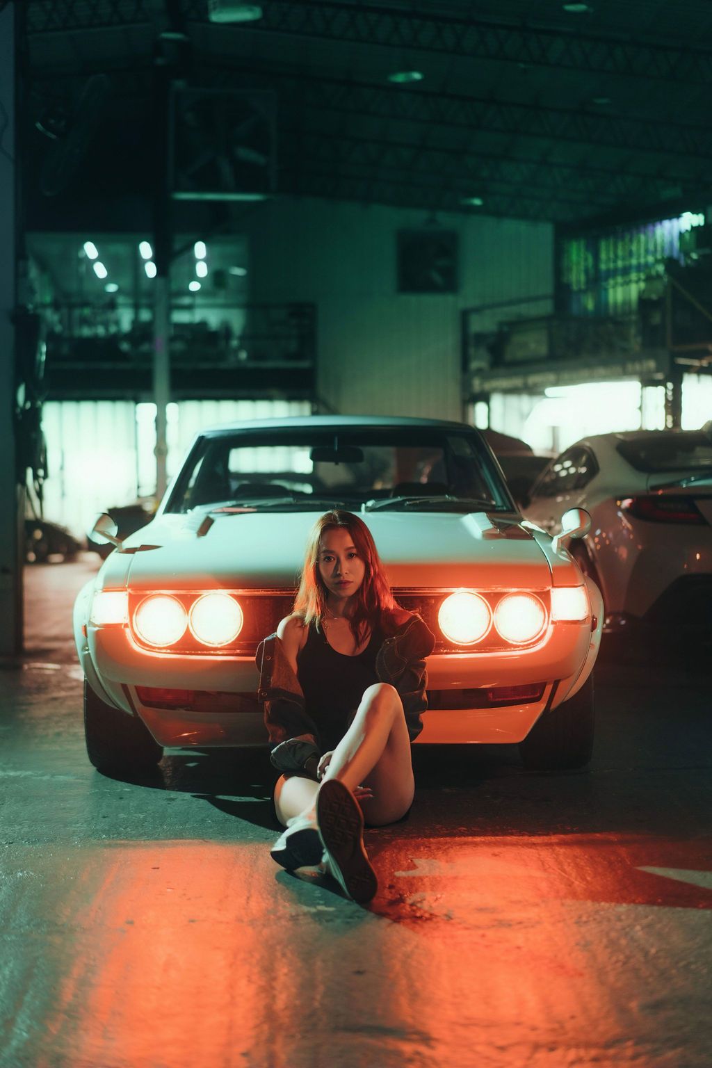 Woman sitting in front of a classic muscle car with glowing headlights in a dimly lit garage at night.
