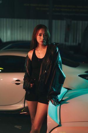 Stylish woman in black outfit and leather jacket poses by white car at night.