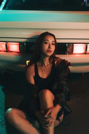 Young woman in black dress and jacket sits against vintage car taillights at night in moody neon lighting.