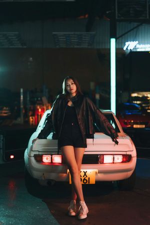 Young woman in leather jacket and shorts leaning on white sports car at night.
