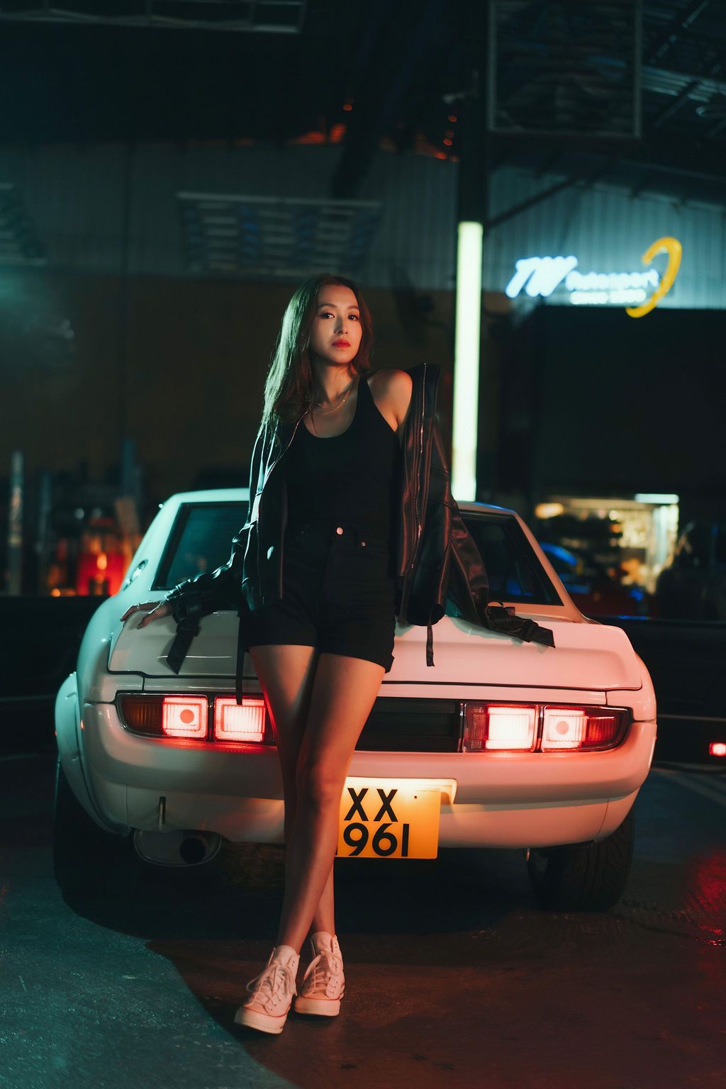 Woman in black shorts and jacket leans on vintage white car under neon lights.