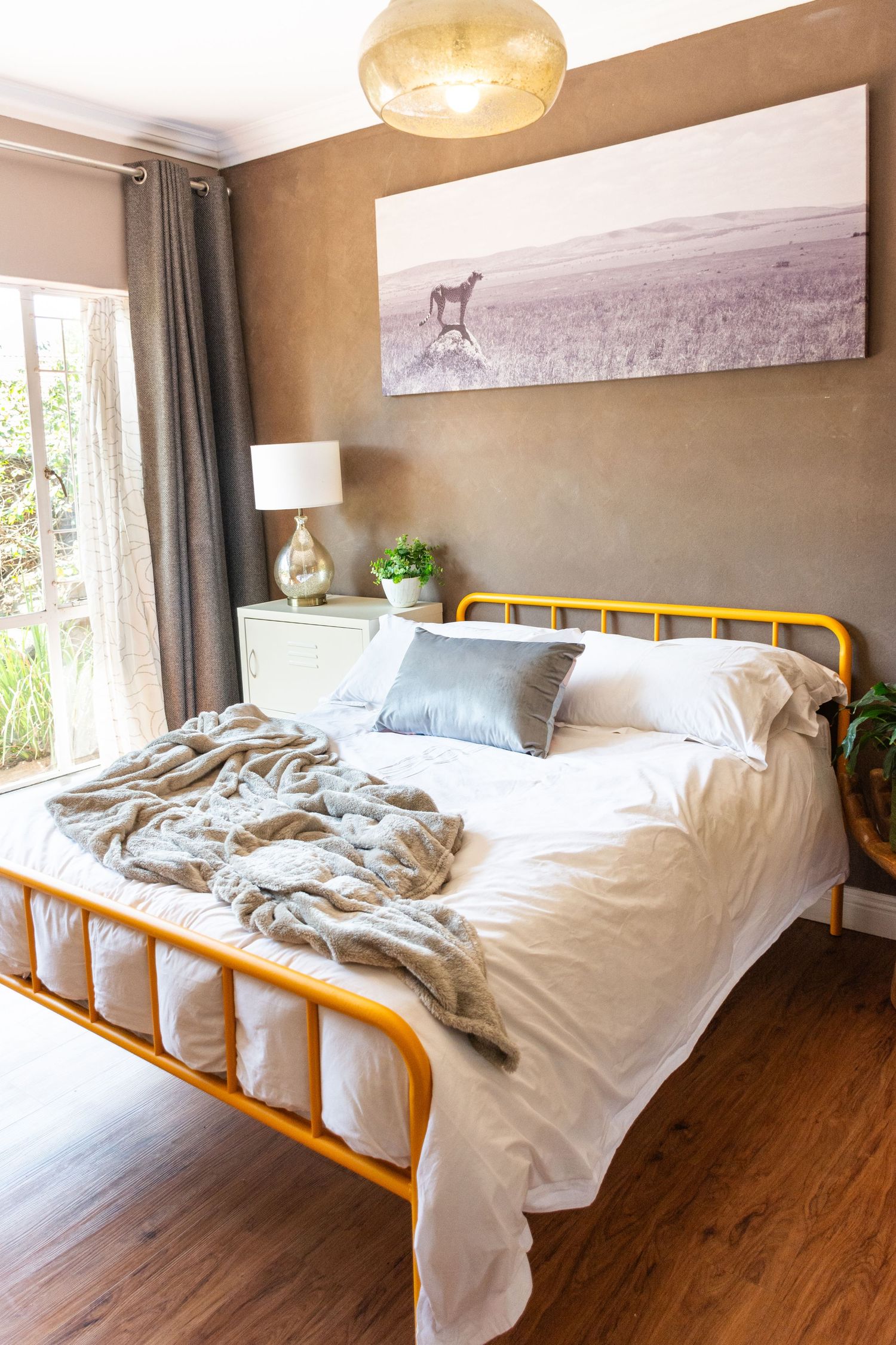 Yellow metal bed frame with white bedding and gray throw in a cozy bedroom with wood floors and safari wall art.