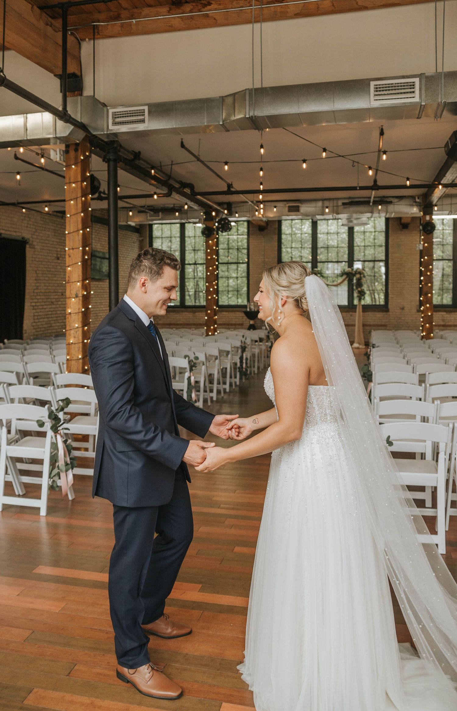 Bride and groom share a first look moment holding hands in a warmly lit industrial wedding venue with string lights.