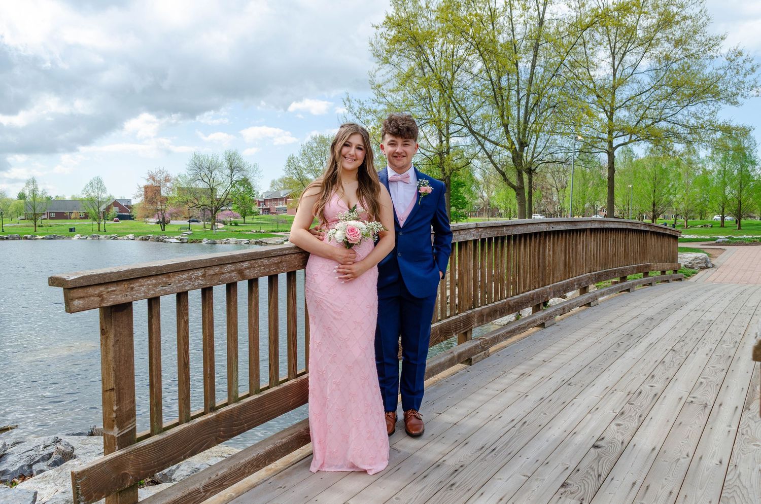 A couple in formal prom attire poses on marble stairs, she in pink gown holding bouquet, he in navy suit.